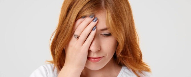 Eine junge Frau mit roten Haaren und weißem T-Shirt hält ihre Hand an ihr rechtes Auge und wirkt nachdenklich oder leicht besorgt. Sie trägt einen silbernen Ring und hat blaue lackierte Fingernägel. Der Hintergrund ist neutral und hell.