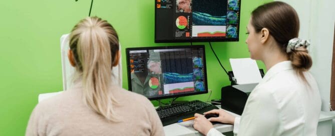 Eine Augenärztin in weißem Kittel sitzt an einem Schreibtisch und analysiert mit einer Patientin eine OCT-Untersuchung (Optische Kohärenztomographie) auf zwei Monitoren. Die Bildschirme zeigen detaillierte Querschnittsbilder der Netzhaut mit farbigen Diagrammen. Die Patientin sitzt mit dem Rücken zur Kamera und trägt einen beigen Pullover. Der Raum hat eine grüne Wand, und im Hintergrund ist medizinische Ausrüstung zu sehen.
